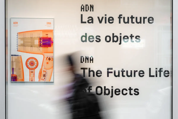 Poster and name of the exhibition behind a busy vitrine. We can read DNA, the future life of objects in French and English. A blurred figure passes through in the foreground.